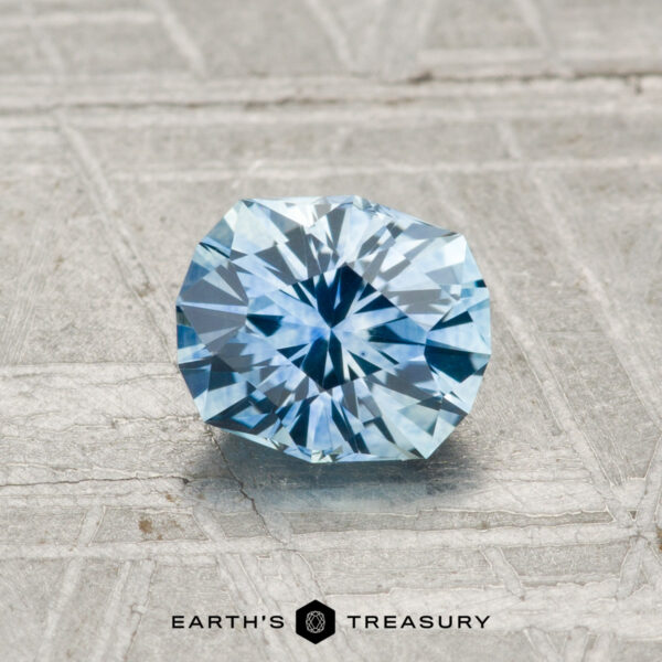 0.96-Carat Montana Sapphire (Heated) A 0.96-carat heated Montana Sapphire, round and faceted in light blue, is displayed on a gray textured surface with "EARTH'S TREASURY" text at the bottom.