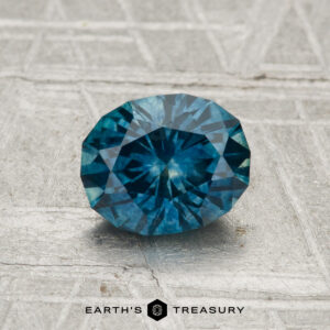A 1.20-carat heated Montana sapphire gemstone sits on a textured gray surface, with "EARTH'S TREASURY" displayed at the bottom.