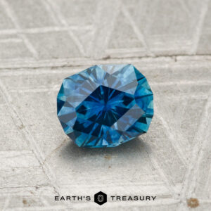 A faceted 0.94-carat heated Montana sapphire with a geometric cut is displayed on a textured gray surface, with the text "EARTH'S TREASURY" at the bottom.
