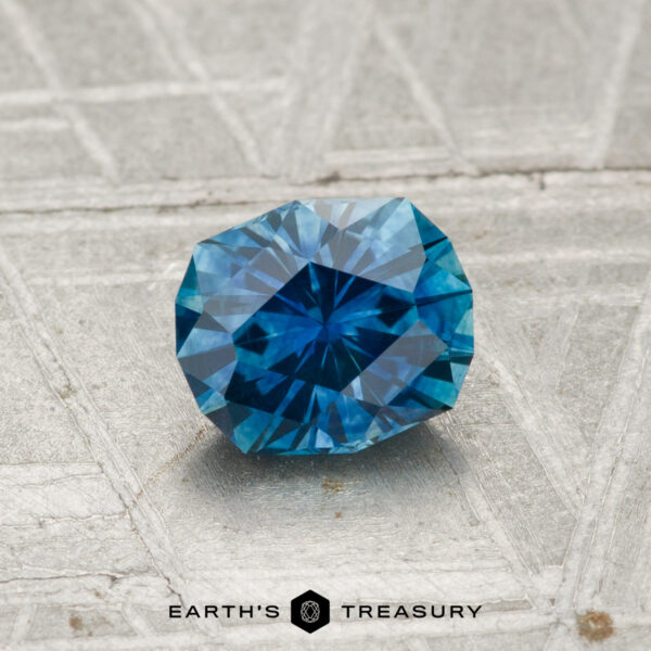 0.94-Carat Montana Sapphire (Heated) A faceted 0.94-carat heated Montana sapphire with a geometric cut is displayed on a textured gray surface, with the text "EARTH'S TREASURY" at the bottom.
