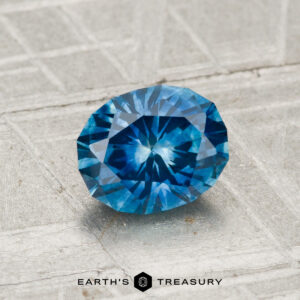 A 1.18-carat heated Montana sapphire is displayed on a textured surface with the "Earth's Treasury" logo and text at the bottom.