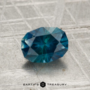 A 1.35-carat Montana Sapphire (heated), oval and blue-green, rests on a textured gray surface with "EARTH'S TREASURY" and a hexagon logo at the bottom.