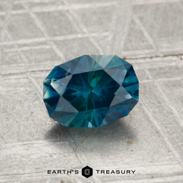 1.35-Carat Montana Sapphire (Heated) A 1.35-carat Montana Sapphire (heated), oval and blue-green, rests on a textured gray surface with "EARTH'S TREASURY" and a hexagon logo at the bottom.
