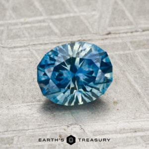 A 1.18-carat heated Montana sapphire, faceted in an oval shape, rests on a textured gray surface with the Earth's Treasury logo at the bottom of the image.