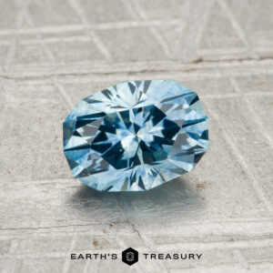 A 1.32-carat heated Montana sapphire sparkles on a textured gray surface, with "EARTH'S TREASURY" and a hexagonal logo displayed at the bottom.