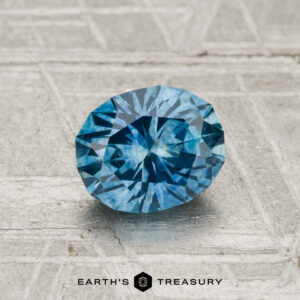 A 1.15-carat heated Montana sapphire with an oval cut is shown on a textured, light surface, featuring the “EARTH’S TREASURY” text and a hexagon logo at the bottom.