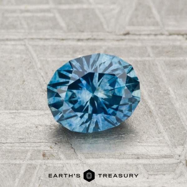 1.15-Carat Montana Sapphire (Heated) A 1.15-carat heated Montana sapphire with an oval cut is shown on a textured, light surface, featuring the “EARTH’S TREASURY” text and a hexagon logo at the bottom.
