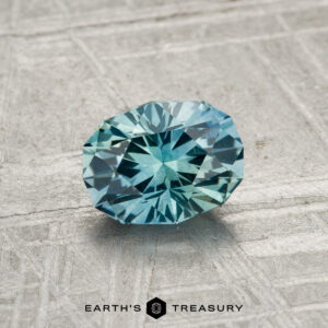 A round, faceted 1.07-carat Montana Sapphire (Heated) with a rich blue hue rests on a textured gray surface, featuring the "EARTH'S TREASURY" text and a hexagon logo at the bottom.