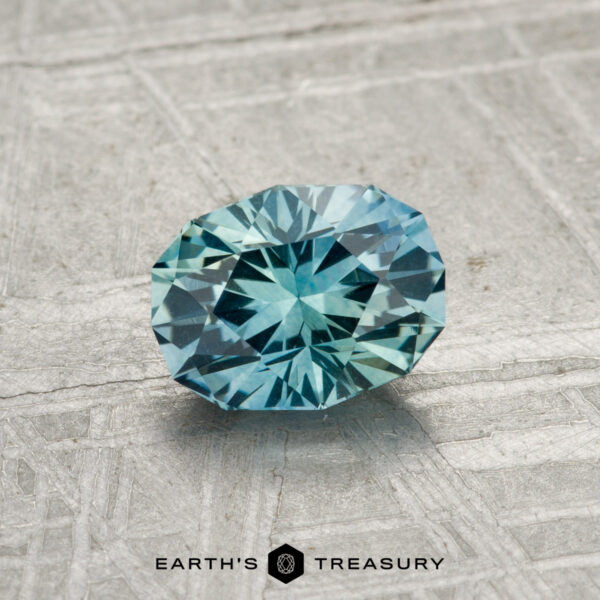 1.07-Carat Montana Sapphire (Heated) A round, faceted 1.07-carat Montana Sapphire (Heated) with a rich blue hue rests on a textured gray surface, featuring the "EARTH'S TREASURY" text and a hexagon logo at the bottom.