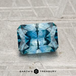 A 1.38-carat heated Montana sapphire, cut in a rectangular teal-blue shape with facets, is displayed on a textured gray surface. "EARTH'S TREASURY" appears at the bottom of the image.