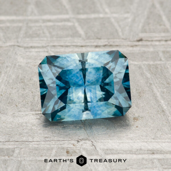 1.38-Carat Montana Sapphire (Heated) A 1.38-carat heated Montana sapphire, cut in a rectangular teal-blue shape with facets, is displayed on a textured gray surface. "EARTH'S TREASURY" appears at the bottom of the image.