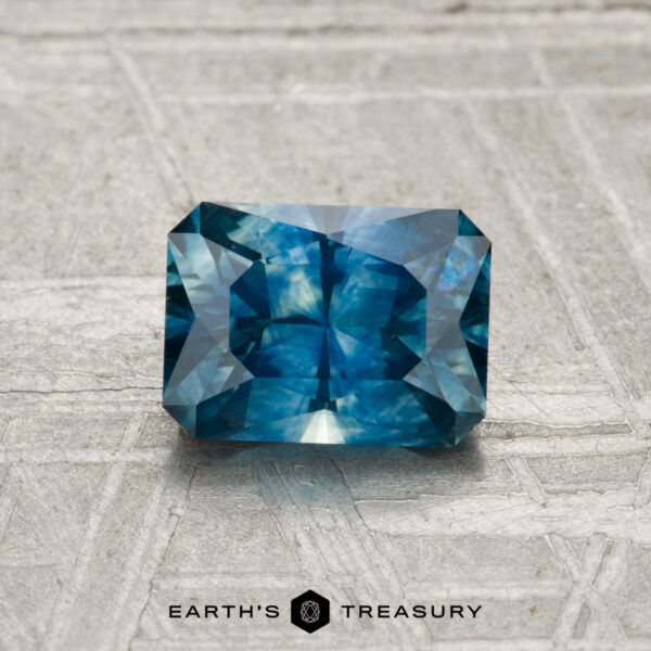 1.22-Carat Montana Sapphire (Heated) A 1.22-carat heated Montana sapphire, rectangular and blue with facets, rests on a textured light gray surface. "EARTH'S TREASURY" text and logo are displayed at the bottom.