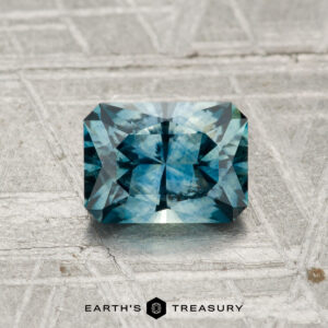 A 1.21-carat heated Montana sapphire, rectangular and blue-green with facets, is displayed on a textured gray surface with "EARTH'S TREASURY" at the bottom.