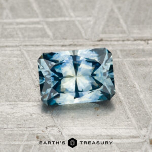 A 1.14-carat heated Montana sapphire with a radiant cut rests on a textured gray surface, featuring the "EARTH'S TREASURY" text and hexagonal logo below.