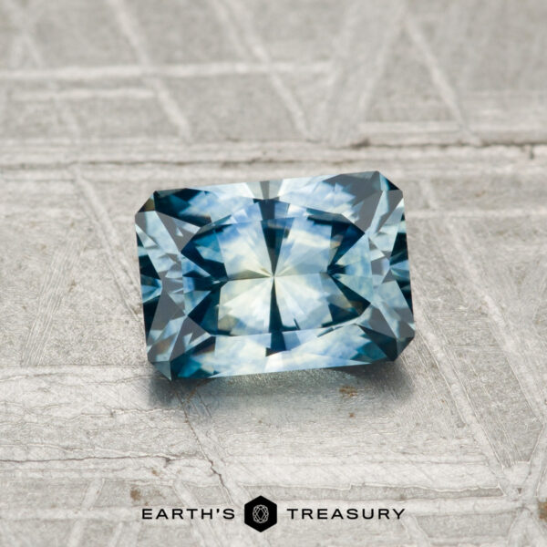 1.14-Carat Montana Sapphire (Heated) A 1.14-carat heated Montana sapphire with a radiant cut rests on a textured gray surface, featuring the "EARTH'S TREASURY" text and hexagonal logo below.