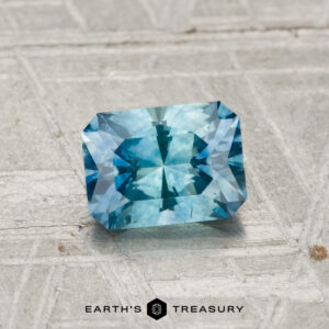 A rectangular, faceted blue 1.10-carat Montana sapphire (heated) rests on a textured gray surface with "EARTH'S TREASURY" displayed below.