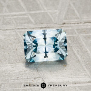 A 1.08-carat heated Montana sapphire, rectangular and faceted in blue, is displayed on a textured gray surface with the "Earth's Treasury" logo at the bottom.