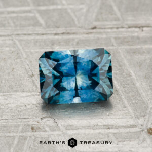 A rectangular, blue-green 1.06-carat Montana sapphire (heated) sits on a textured gray surface with the text "EARTH'S TREASURY" at the bottom.