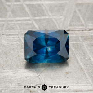 Rectangular blue 1.06-carat heated Montana sapphire with multiple facets, showcased on a textured grey surface. "Earth’s Treasury" logo is visible at the bottom of the image.