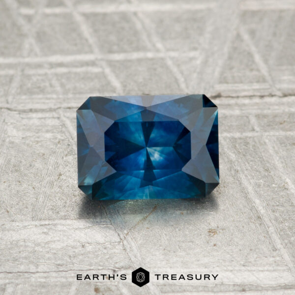 1.06-Carat Montana Sapphire (Heated) Rectangular blue 1.06-carat heated Montana sapphire with multiple facets, showcased on a textured grey surface. "Earth’s Treasury" logo is visible at the bottom of the image.