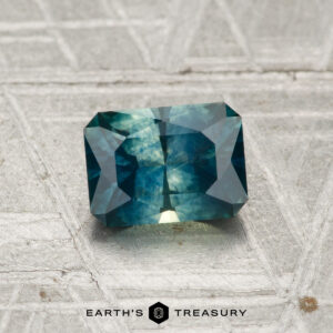 A 1.03-carat heated Montana sapphire with a radiant cut and blue-green hue is shown on a textured silver-gray surface, with "EARTH'S TREASURY" at the bottom.