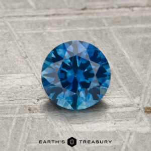 A 1.03-carat heated Montana sapphire with a round brilliant cut rests on a textured gray surface. The EARTH'S TREASURY logo appears at the bottom.