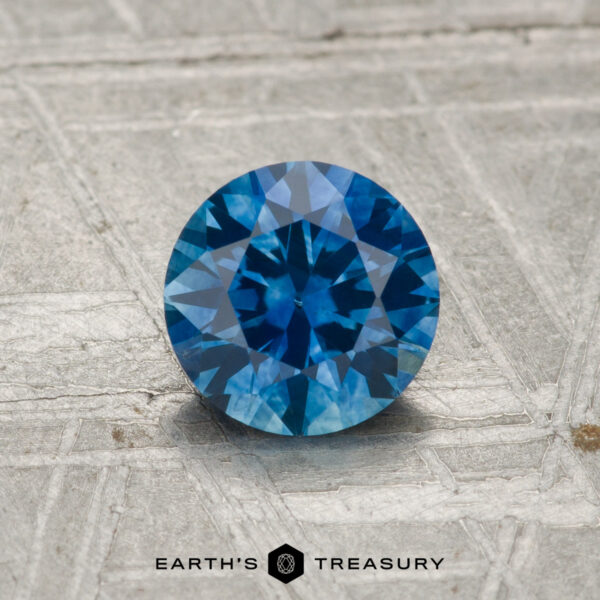 1.03-Carat Montana Sapphire (Heated) A 1.03-carat heated Montana sapphire with a round brilliant cut rests on a textured gray surface. The EARTH'S TREASURY logo appears at the bottom.