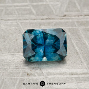 A 1.02-carat heated Montana sapphire, cut in a rectangular blue-green facet, is displayed on a gray patterned surface with "EARTH'S TREASURY" at the bottom.