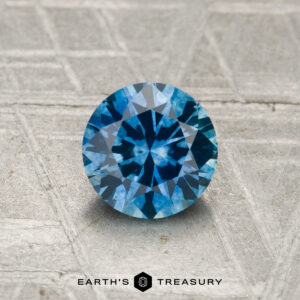 A 1.01-carat heated Montana sapphire, featuring a round brilliant cut, is displayed on a textured surface with "EARTH'S TREASURY" written below.