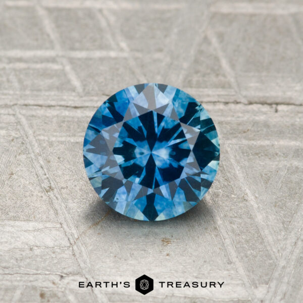 1.01-Carat Montana Sapphire (Heated) A 1.01-carat heated Montana sapphire, featuring a round brilliant cut, is displayed on a textured surface with "EARTH'S TREASURY" written below.