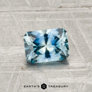 A 0.98-carat heated Montana sapphire, cut in a rectangular faceted shape, rests on a textured gray surface. The text "Earth's Treasury" appears at the bottom of the image.