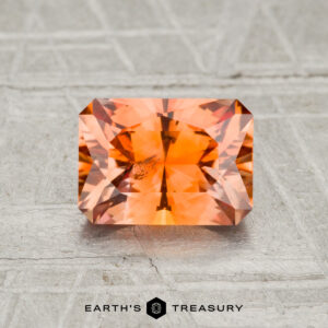 A rectangular, faceted 1.52-carat Montana sapphire is displayed on a light textured surface with the text “Earth’s Treasury” below.