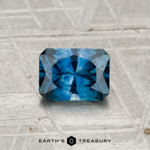 A 0.98-carat heated Montana sapphire with intricate blue facets is displayed on a textured light gray surface. "EARTH'S TREASURY" appears at the bottom of the image.