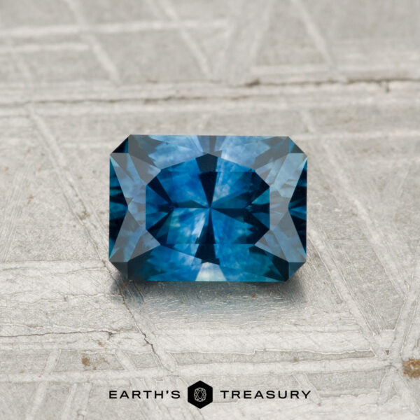 0.98-Carat Montana Sapphire (Heated) A 0.98-carat heated Montana sapphire with intricate blue facets is displayed on a textured light gray surface. "EARTH'S TREASURY" appears at the bottom of the image.
