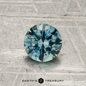 A 1.00-carat heated Montana sapphire, round and faceted, rests on a textured metallic surface with "Earth's Treasury" displayed at the bottom center.