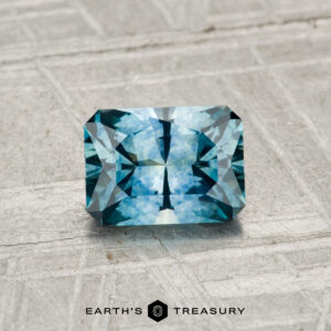 A 0.96-carat rectangular blue Montana sapphire (heated) with intricate facets rests on a textured light gray surface. The Earth's Treasury logo appears at the bottom of the image.