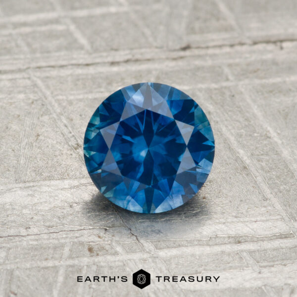 0.99-Carat Montana Sapphire (Heated) A 0.99-carat round-cut heated Montana sapphire shown on a textured gray surface, with "EARTH'S TREASURY" at the bottom.