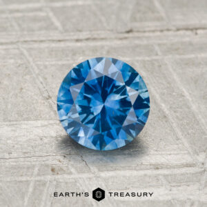 A 0.99-carat heated Montana sapphire with a round brilliant cut sits on a textured metallic surface, with the Earth’s Treasury logo visible at the bottom.
