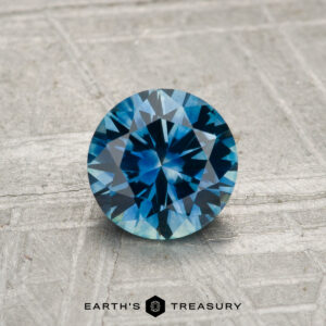 A 0.99-carat round-cut heated Montana sapphire is displayed on a textured gray surface, with "Earth's Treasury" shown at the bottom.