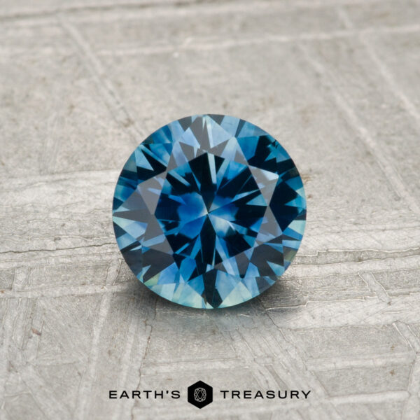 0.99-Carat Montana Sapphire (Heated) A 0.99-carat round-cut heated Montana sapphire is displayed on a textured gray surface, with "Earth's Treasury" shown at the bottom.
