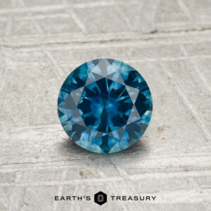 A 0.95-carat heated Montana sapphire, round and faceted, rests on a textured metallic surface with "EARTH'S TREASURY" displayed at the bottom.