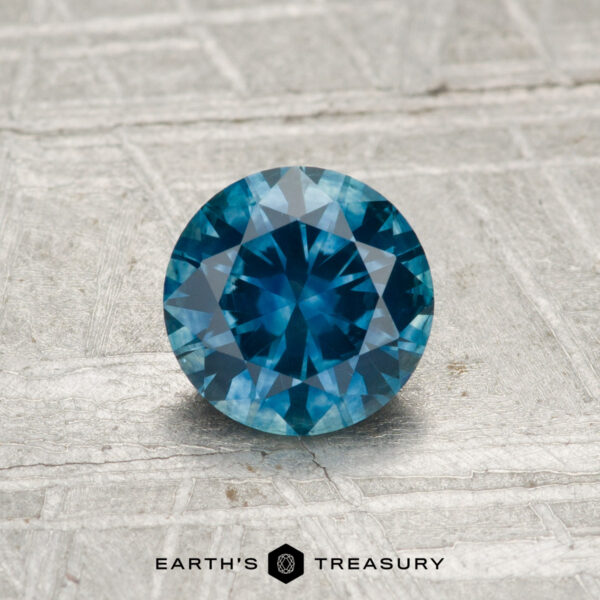 0.95-Carat Montana Sapphire (Heated) A 0.95-carat heated Montana sapphire, round and faceted, rests on a textured metallic surface with "EARTH'S TREASURY" displayed at the bottom.