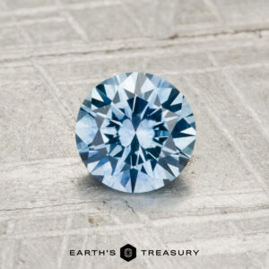 A 0.93-carat Montana Sapphire (heated), round brilliant-cut and blue, is showcased on a textured metallic surface with "EARTH'S TREASURY" at the bottom.