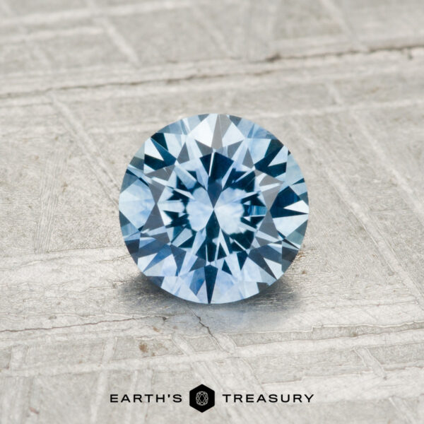 0.93-Carat Montana Sapphire (Heated) A 0.93-carat Montana Sapphire (heated), round brilliant-cut and blue, is showcased on a textured metallic surface with "EARTH'S TREASURY" at the bottom.