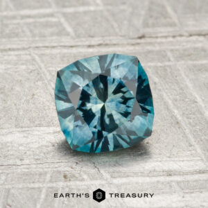 A 1.19-carat heated Montana sapphire, expertly square-cut with intricate facets, rests on a textured gray surface. “EARTH’S TREASURY” is displayed at the bottom.