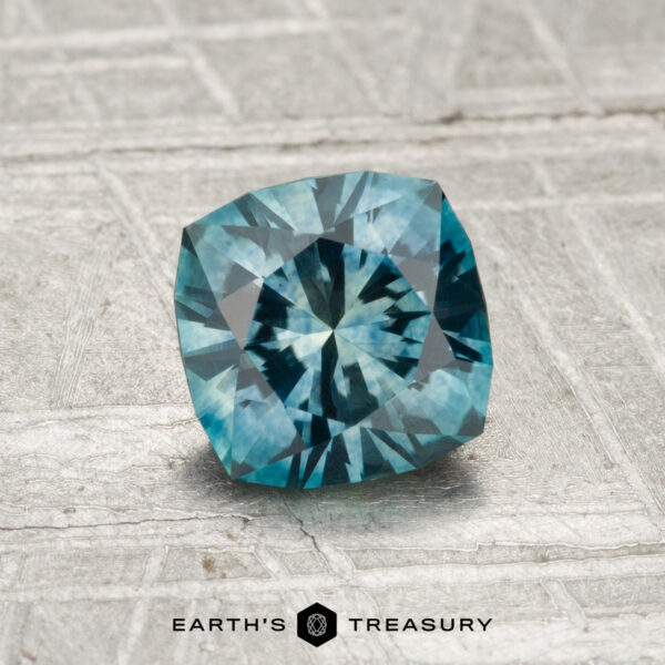 1.19-Carat Montana Sapphire (Heated) A 1.19-carat heated Montana sapphire, expertly square-cut with intricate facets, rests on a textured gray surface. “EARTH’S TREASURY” is displayed at the bottom.