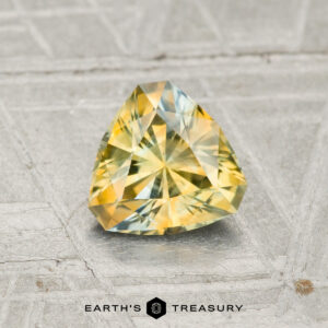 A faceted, triangular 0.95-carat Montana Sapphire gemstone is displayed on a textured gray surface with the text "EARTH'S TREASURY" below.