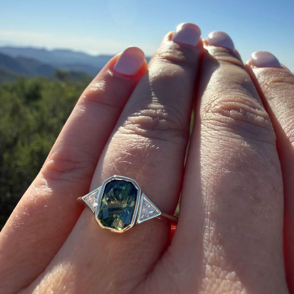 A hand with light pink nails displays the Cathedral "Ceres" ring in 14K yellow gold, set with a 1.52-carat Australian sapphire, along with two triangular clear stones. The background shows a blurred outdoor landscape.