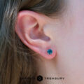 Close-up of a person's ear wearing a 5.25 teal heated Montana Sapphire earring; part of their brown hair is visible. Earth's Treasury logo appears at the bottom.