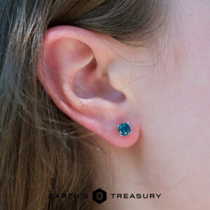 Close-up of a person's ear wearing a 5.25 teal heated Montana Sapphire earring; part of their brown hair is visible. Earth's Treasury logo appears at the bottom.